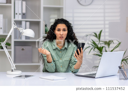 Perplexed mature Latina professional at desk with laptop and mobile phone, managing work tasks in a bright home office. Perplexed mature Latina professional at desk with laptop and mobile phone, managing work tasks in a bright home office. 111537452