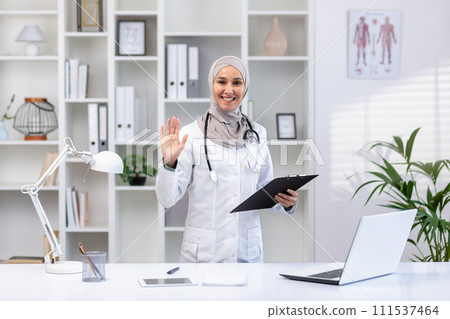 Smiling Muslim doctor in hijab greeting with a wave while holding a clipboard in a well-lit medical office. Smiling Muslim doctor in hijab greeting with a wave while holding a clipboard in a well-lit medical office. 111537464