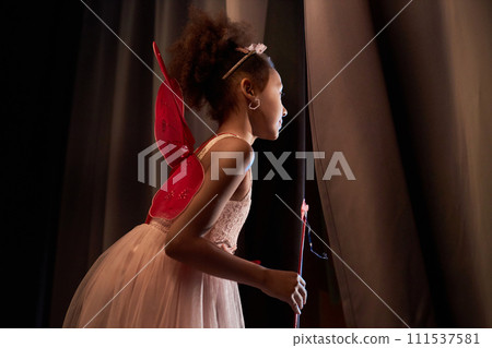 Side view young Black girl wearing fairy costume with wings peeking over curtain backstage in theater copy space 111537581