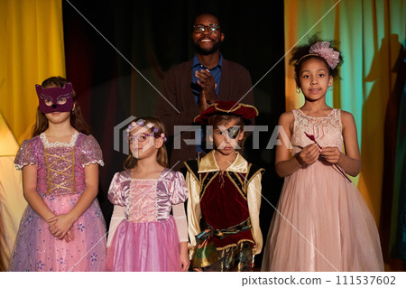 Front view at group of children actors on stage in theater with smiling drama teacher applauding 111537602