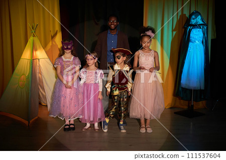 Full length portrait of group of little kids standing on stage in theater with smiling drama teacher Full length portrait of group of little kids standing on stage in theater with smiling drama teacher 111537604