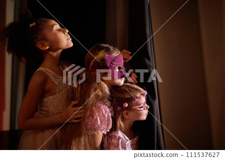 Side view portrait of three little girls wearing costumes peeking over curtain backstage in theater copy space 111537627