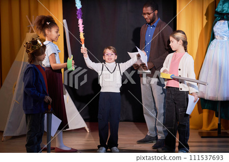 Full length portrait of young girl standing on stage rehearsing school play with group of children in theater Full length portrait of young girl standing on stage rehearsing school play with group of children in theater 111537693