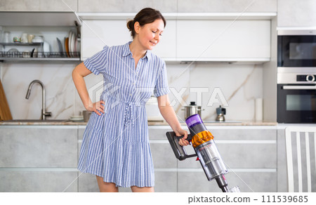 Smiling housewife in blue dress run vacuum cleaner floor covering near dining table Smiling housewife in blue dress run vacuum cleaner floor covering near dining table 111539685