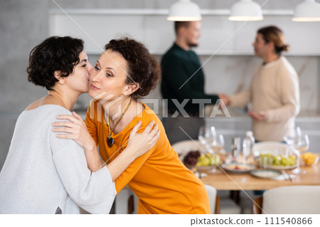 Women hug each other and kiss on the cheek while greeting each other before a festive dinner. Men shaking hands in background 111540866