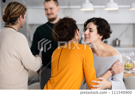 Women hug each other and kiss on the cheek while greeting each other before a festive dinner. Men shaking hands in background 111540900