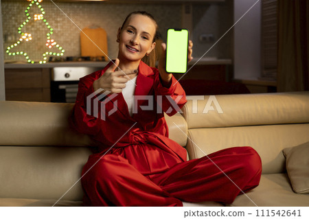 A young woman in red pajamas shows a green smartphone screen in a cozy living room during Christmas 111542641
