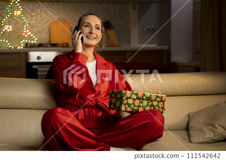Smiling Woman in Red Pajamas Talking on Phone With Christmas Gift on Couch at Home 111542642