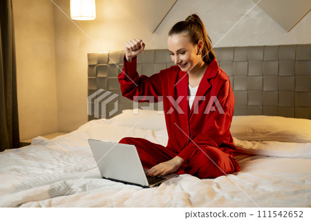 Excited Woman Celebrates Online Win in Red Pajamas Sitting on Bed at Home Excited Woman Celebrates Online Win in Red Pajamas Sitting on Bed at Home 111542652