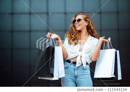 Happy stylish woman with shopping bags enjoys sunny weather walking down street. Summer shopping. Happy stylish woman with shopping bags enjoys sunny weather walking down street. Summer shopping. 111543134