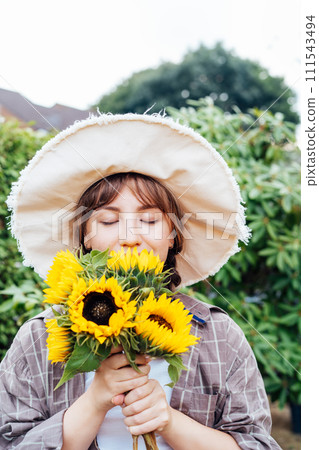 Dreaming young female farmer woman with closed eyes holding and sniffing a sunflowers bouquet on the green garden background. Enjoy the moment. Rural, Cottage core lifestyle. Vertical card 111543494