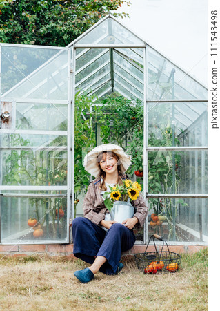 Portrait of smiling young female farmer woman holding watering can with fresh sunflowers bouquet, sitting near greenhouse. Cottagecore lifestyle. Growing organic vegetables in garden. Vertical card. Portrait of smiling young female farmer woman holding watering can with fresh sunflowers bouquet, sitting near greenhouse. Cottagecore lifestyle. Growing organic vegetables in garden. Vertical card. 111543498