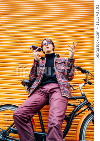 Young hipster man recording voice message on phone while sitting on cruiser bicycle near orange wall. Student using 5g network. Eco friendly transport for carbon footprint. Sustainable lifestyle. Young hipster man recording voice message on phone while sitting on cruiser bicycle near orange wall. Student using 5g network. Eco friendly transport for carbon footprint. Sustainable lifestyle. 111543503