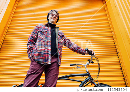 Bottom view Young stylish hipster man standing with a cruiser city Bicycle near orange Wall. Student Resting after ride bicycle. Eco friendly transportation for carbon footprint. Sustainable lifestyle Bottom view Young stylish hipster man standing with a cruiser city Bicycle near orange Wall. Student Resting after ride bicycle. Eco friendly transportation for carbon footprint. Sustainable lifestyle 111543504