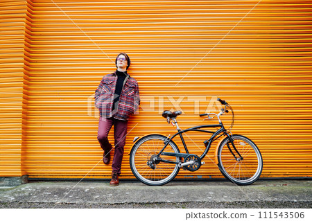 Young stylish hipster man standing with a cruiser city Bicycle near orange Wall. Student Resting after ride bicycle. Eco friendly transportation for carbon footprint. Sustainable lifestyle 111543506