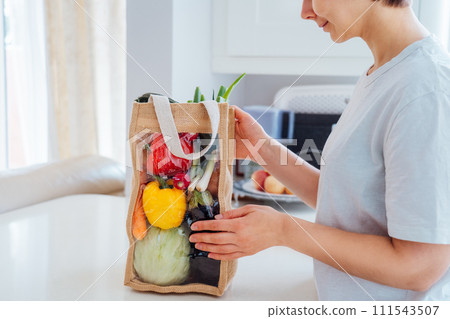 Woman with reusable eco sackcloth fabric bag packaging with fresh organic vegetables on white kitchen table. Home food delivery concept. Local farmer healthy food. Eco-friendly, Sustainable lifestyle. Woman with reusable eco sackcloth fabric bag packaging with fresh organic vegetables on white kitchen table. Home food delivery concept. Local farmer healthy food. Eco-friendly, Sustainable lifestyle. 111543507