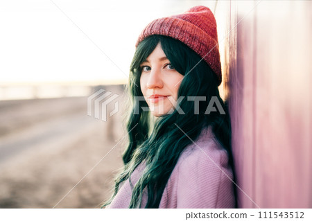 Portrait of relaxed stylish young smiling hipster woman with color hair wearing pink coat and knitted hat leaning on beach hut and enjoying moment. Simple pleasures and personal fulfillment. 111543512