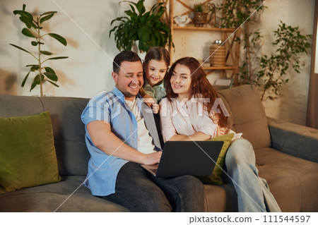 Choosing destination via Internet. Happy family, mother, father and daughter sitting at home, looking on laptop and planning upcoming trip 111544597