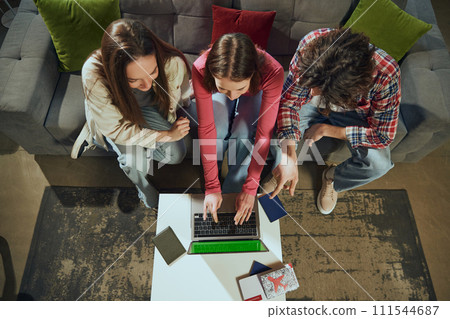 Top view image of young boy and girls sitting on couch at home and using laptop for booking tickets, making hotel reservation. Going on vacation Top view image of young boy and girls sitting on couch at home and using laptop for booking tickets, making hotel reservation. Going on vacation 111544687
