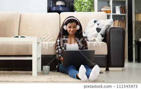 A dark-skinned girl in headphones with laptop on floor at home A dark-skinned girl in headphones with laptop on floor at home 111545973