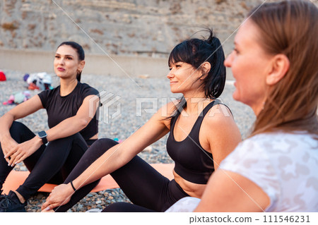 Group of smiling young Caucasian women are sitting on sports mats on wild beach and talking to each other. Concept of female circle of communication and female support Group of smiling young Caucasian women are sitting on sports mats on wild beach and talking to each other. Concept of female circle of communication and female support 111546231