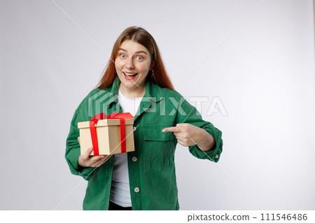 Portrait of a smiling casual woman holding present and pointing finger over white background. Happy woman receive gift, smiling excited. Valentine's Day Women's Day birthday holiday concept. Portrait of a smiling casual woman holding present and pointing finger over white background. Happy woman receive gift, smiling excited. Valentine's Day Women's Day birthday holiday concept. 111546486