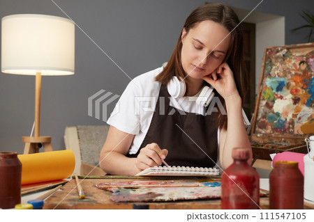 Artistic inspiration in painting. Professional artist's creative workshop. Caucasian brown haired woman artist drawing with paints sitting in studio workshop 111547105