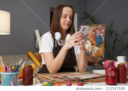 Professional painter's exhibition success. Adorable brown haired woman artist drawing with paints and using mobile phone sitting in studio workshop browsing internet pages while having break 111547109
