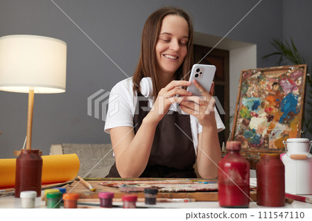 Smiling satisfied Caucasian woman wearing brown apron painting in art creative studio and using smartphone scrolling online searching ideas for pictures 111547110
