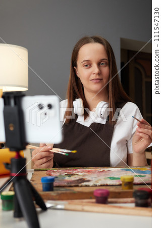 Brown haired woman painter blogger wearing brown apron painting using mobile phone on tripod at her workplace broadcasting livestream online drawing lesson 111547130