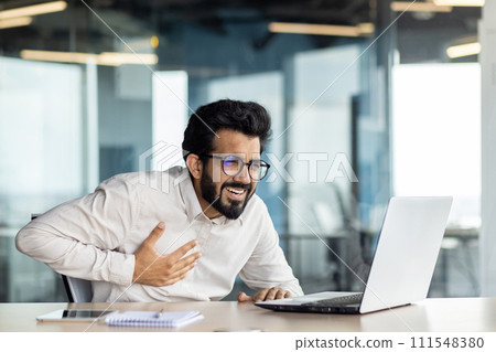 Heart attack at work. A young Indian man is sitting at the desk in the office and holding his hands to his chest, feeling fear and severe pain. 111548380