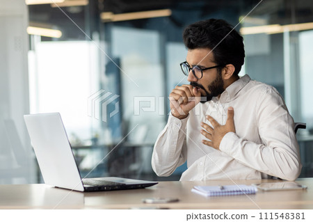 A young Indian man came down with a virus and a cold. Sitting in the office at the table and coughing, holding his chest from pain and covering his mouth with his hand. 111548381