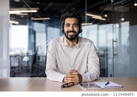Portrait of a smiling Muslim man businessman, marketer, salesman sitting in the office at the table in a headset and smiling confidently at the camera. Portrait of a smiling Muslim man businessman, marketer, salesman sitting in the office at the table in a headset and smiling confidently at the camera. 111548405