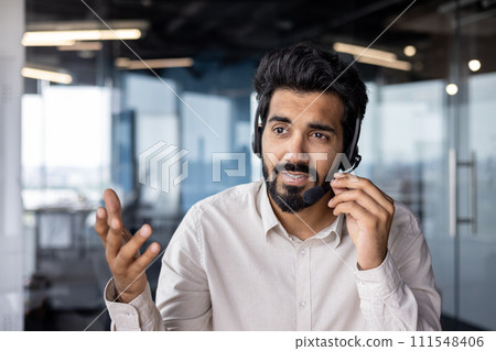 Close-up photo of a young Indian male businessman wearing a headset is in the office and talking and consulting through a microphone, gesturing with his hands. Close-up photo of a young Indian male businessman wearing a headset is in the office and talking and consulting through a microphone, gesturing with his hands. 111548406