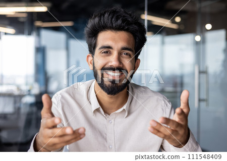 Close-up portrait of smiling young male businessman, office worker looking at camera, talking on video call and gesturing with hands. Close-up portrait of smiling young male businessman, office worker looking at camera, talking on video call and gesturing with hands. 111548409