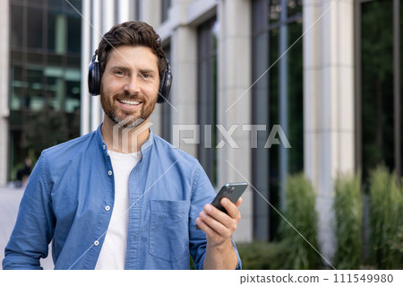 A smiling bearded businessman in casual attire enjoying music on headphones while using a smartphone, standing near an office building. A smiling bearded businessman in casual attire enjoying music on headphones while using a smartphone, standing near an office building. 111549980