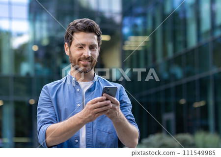 Casual mature businessman with a beard happily using a smartphone outside a glass office building, portraying a relaxed professional. 111549985