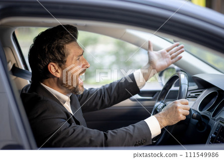 Close-up photo of angry young businessman in suit driving car and emotionally gesturing with hands, displeased shouting and arguing. Close-up photo of angry young businessman in suit driving car and emotionally gesturing with hands, displeased shouting and arguing. 111549986