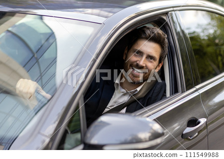 Close-up portrait of a young handsome and successful businessman in a suit sitting behind the wheel of an expensive car, looking out the window and smiling at the camera. 111549988