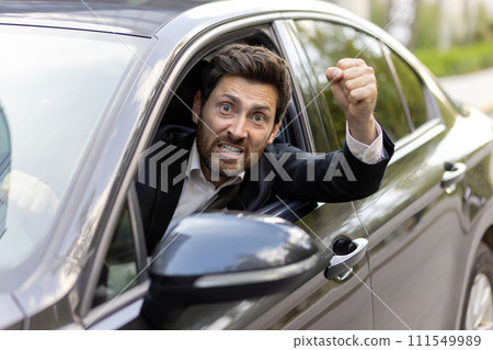 An angry and irritated young man in a business suit looks out of the car window. He is behind the wheel, shouting at the camera and waving his hands threateningly. Close-up photo. 111549989