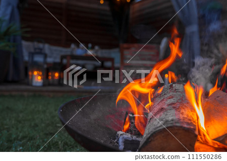 Evening ambiance with a lively fire in a metal pit, casting a warm glow in a garden with a wooden cabin or bar in the background, possibly in Copenhagen. 111550286