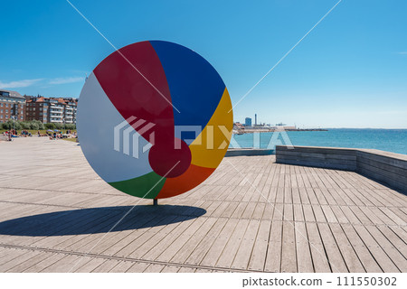 A vibrant circular sculpture with six colored segments stands on a wooden boardwalk by the sea, with a sandy beach and urban backdrop, likely in Helsingborg. A vibrant circular sculpture with six colored segments stands on a wooden boardwalk by the sea, with a sandy beach and urban backdrop, likely in Helsingborg. 111550302