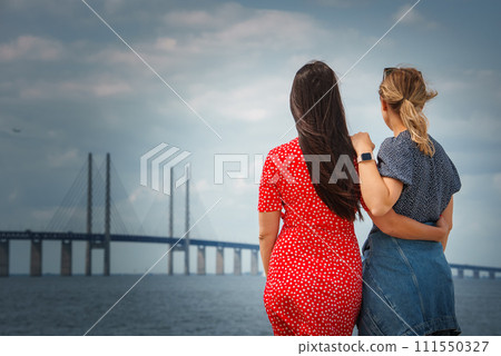 Two women gaze at the Oresund Bridge from Denmark to Sweden, one in a polkadotted red dress, the other in a blue outfit. Overcast skies hint at evening serenity. Two women gaze at the Oresund Bridge from Denmark to Sweden, one in a polkadotted red dress, the other in a blue outfit. Overcast skies hint at evening serenity. 111550327