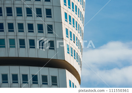 Closeup of a modern curved building with a pattern of white panels and bluetinted windows, accented by a brown horizontal band, against a partly cloudy sky, likely in Malmo. Closeup of a modern curved building with a pattern of white panels and bluetinted windows, accented by a brown horizontal band, against a partly cloudy sky, likely in Malmo. 111550361