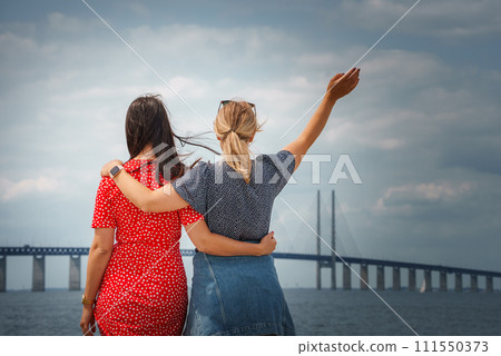 Two friends admire the Oresund Bridge connecting Copenhagen and Malmo, one in a red polka dot dress, the other in a denim skirt and blouse, under a partly cloudy sky. Two friends admire the Oresund Bridge connecting Copenhagen and Malmo, one in a red polka dot dress, the other in a denim skirt and blouse, under a partly cloudy sky. 111550373