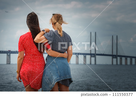 Two friends share a casual embrace by the water, one in a red polka dot dress, the other in a blue outfit, gazing at the Oresund Bridge under a dramatic sky. 111550377