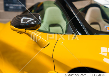 Closeup of a vibrant yellow McLaren 765LT with a carbon fiber side mirror and sleek bodywork, slightly open door revealing a light interior, in a blurred indoor setting. 111550380