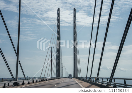 Modern Oresund Bridge connecting Copenhagen and Malmo, viewed from the deck with tall pylons, multiple cables, lane markings, and safety barriers under a partly cloudy sky. Modern Oresund Bridge connecting Copenhagen and Malmo, viewed from the deck with tall pylons, multiple cables, lane markings, and safety barriers under a partly cloudy sky. 111550384