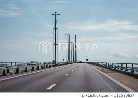 Modern cablestayed bridge under a clear sky, with white lane markings and metal railings, overlooking a calm sea, likely the Oresund Bridge connecting Denmark and Sweden. 111550624