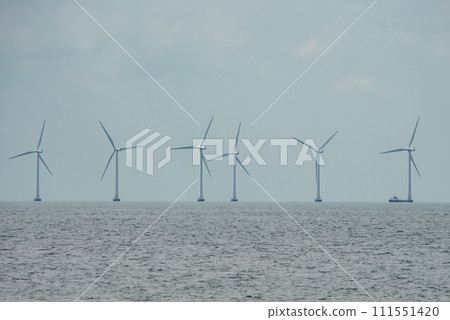 Large wind turbines rotate in the calm sea under an overcast sky, lined up towards the horizon in the Oresund Strait between Copenhagen and Malmo, with a boat near the last turbine for scale. 111551420